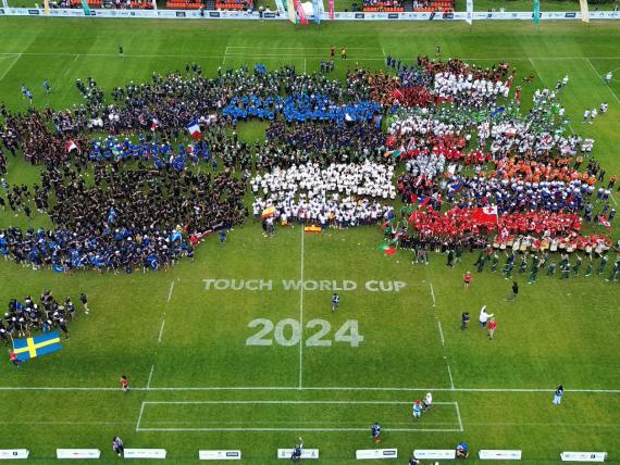 Overhead shot of the rugby pitch with teems assembled and showing the 2024 Touch world Cup logo stencilled on the grass